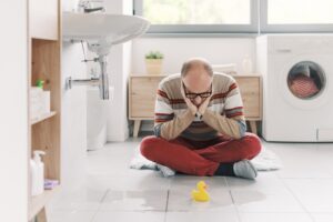 A man sitting on a bathroom floor looking at a puddle from a common plumbing issue known as water leak.