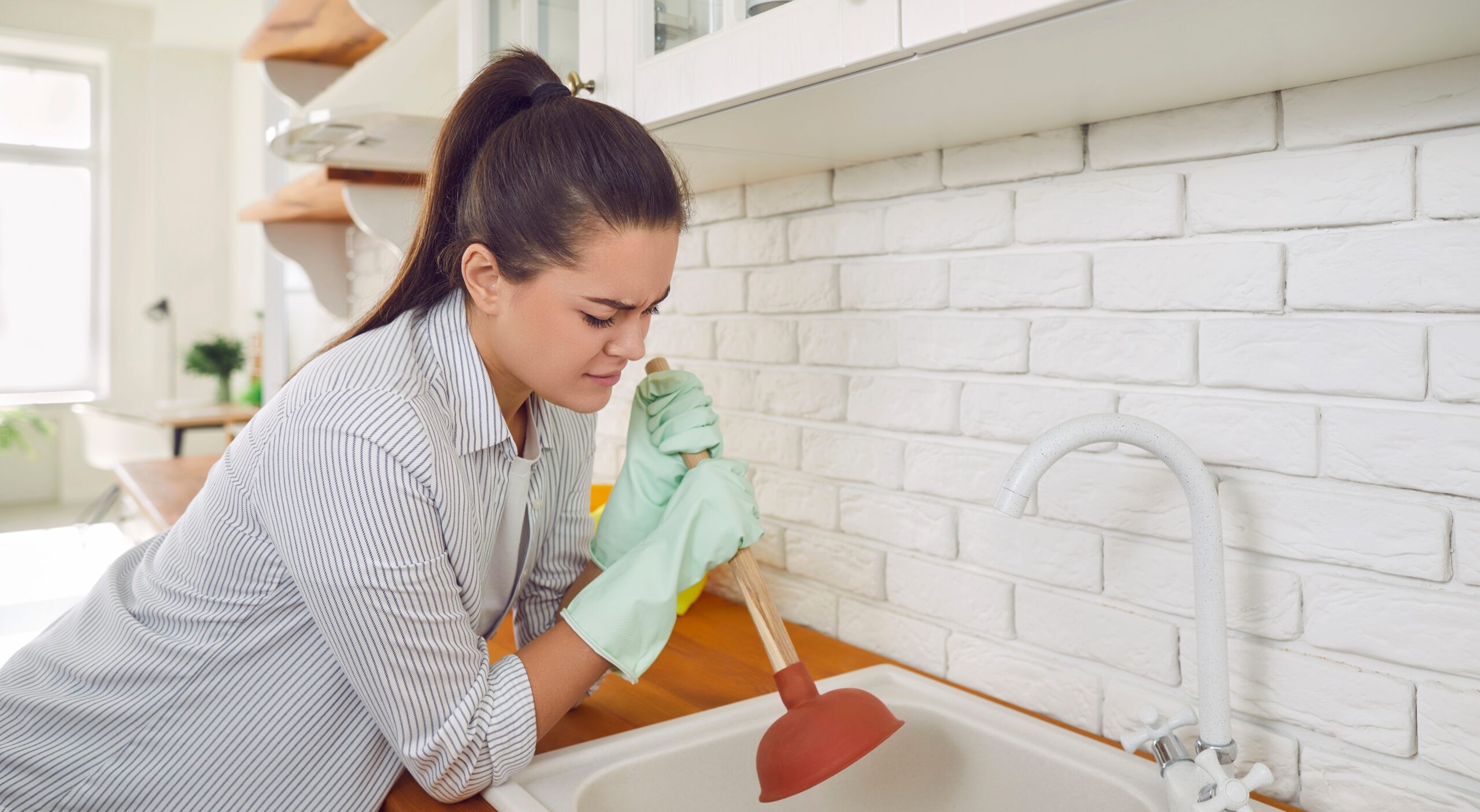 Frustrated homeowner plunging a clogged sink drain in a bright kitchen